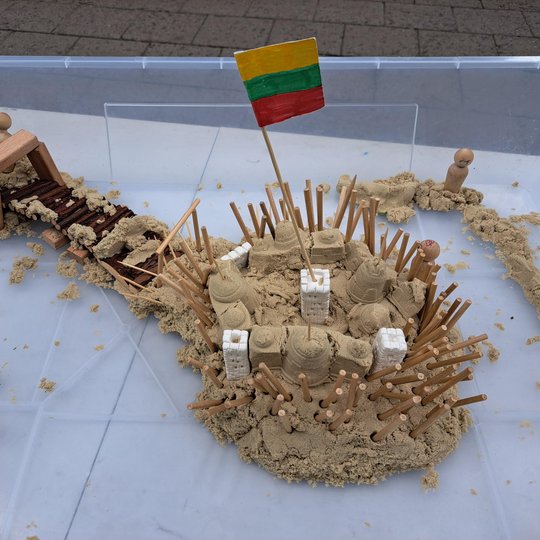 Nuotraukoje matyti vaikų sukurta piliakalnio maketo instaliacija. Ji pagaminta iš smėlio, medinių pagaliukų, putplasčio detalių bei kitų kūrybinių priemonių. Maketas vaizduoja įtvirtintą piliakalnį su vartais, tiltu, sargybiniais bei Lietuvos vėliava cent
