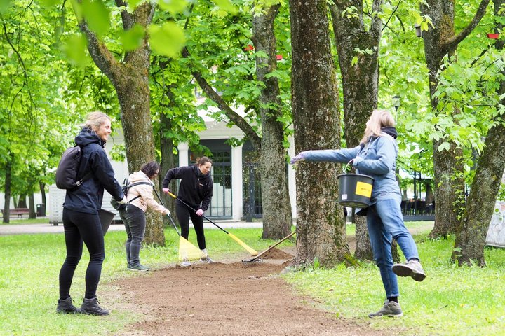 Grupė žmonių parke tvarko žemės taką, naudodami grėblius ir kibirus. Aplink juos žaliuoja medžiai ir matyti balto pastato fragmentas fone.