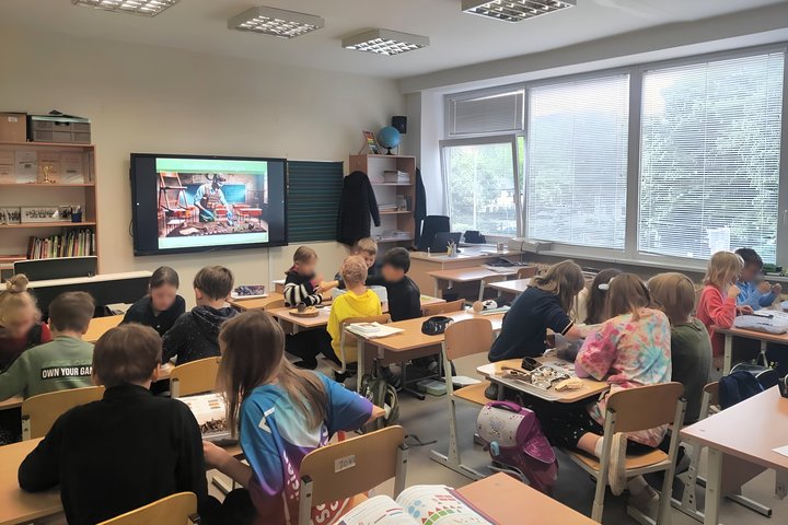 Pradinės mokyklos klasėje grupė vaikų sėdi prie stalų ir dirba komandose. Jie žiūri į įvairius daiktus ant stalų. Klasėje šviesu, pro langus matyti žaluma, o atmosfera atrodo aktyvi ir kūrybiška.