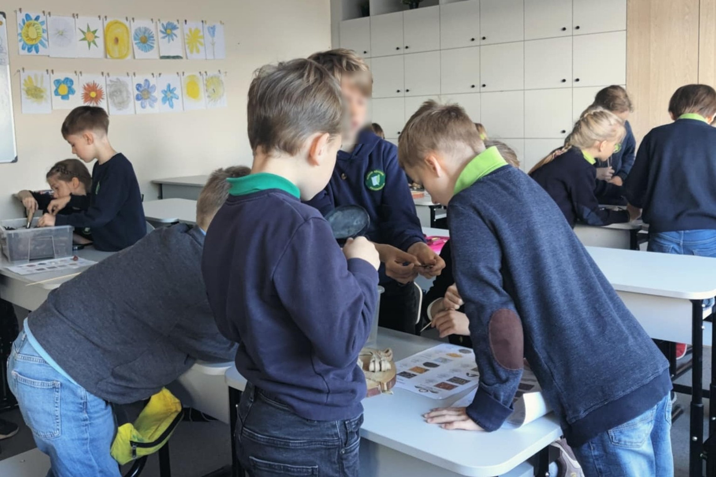 Pradinukai dalyvauja archeologinėje edukacijoje, kuri vyksta klasėje. Vaikai, apsirengę mokyklinėmis uniformomis su tamsiai mėlynais megztiniais ir žaliomis apykaklėmis, susibūrę grupėmis prie stalų, tyrinėja radinių imitacijas – apžiūrinėja šukes ir kitu