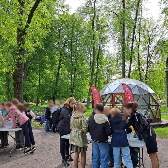 Vaikai susibūrę prie kelių stalų dalyvauja edukacinėje veikloje lauke, apsupti vešlios žalumos. Fone matyti stiklinis kupolas su dviem raudonomis vėliavomis. Vaikus stebi ir padeda kelios mokytojos. Aplinka – pavasariškai žaliuojantis parkas su suoliukais