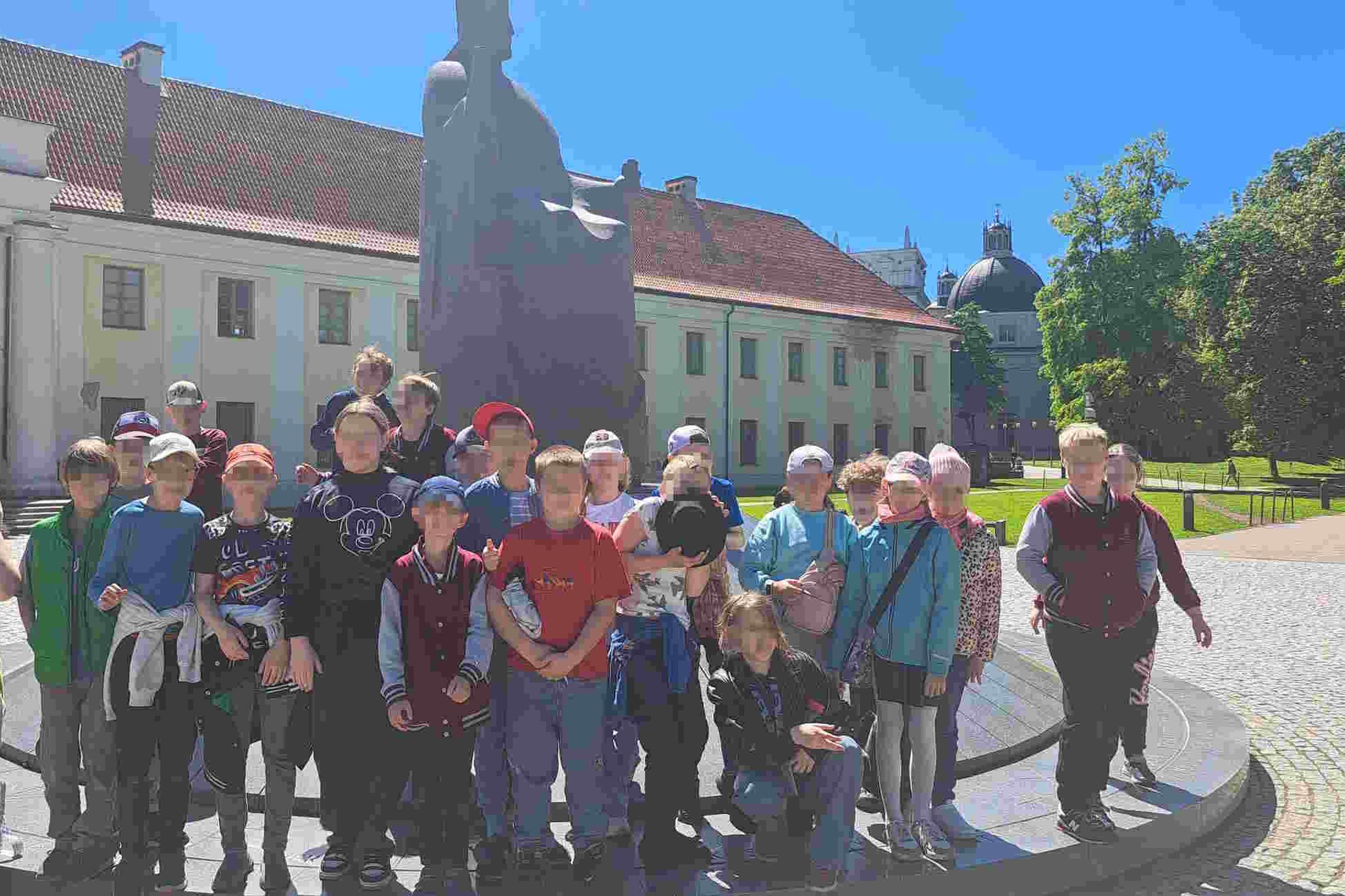 Grupė trečiokų pozuoja prie kunigaikščio Gedimino paminklo Nacionalinio muziejaus Vilniuje kieme, saulėtą dieną.