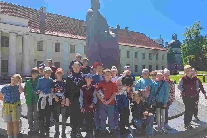 Grupė trečiokų pozuoja prie kunigaikščio Gedimino paminklo Nacionalinio muziejaus Vilniuje kieme, saulėtą dieną.