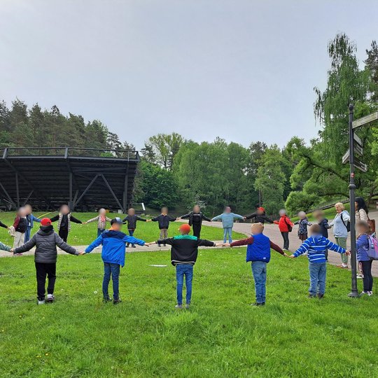 nuotraukoje matome vaikų grupę, susikibusią rankomis ir sudarančią ratą žalioje pievoje prie miško šlaito. Visi vaikai vilki ryškias geltonas liemenes – tai rodo jų dalyvavimą organizuotoje veikloje. Juos lydi mokytoja. Aplinka žalia, gyva ir saugi mokymu