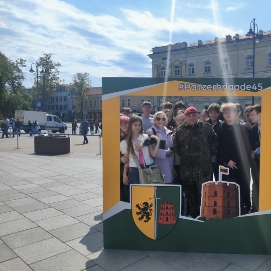 Grupė paauglių mokinių ir jų mokytoja pozuoja nuotraukai kartu su Vokietijos kariu, vilkinčiu maskuojamąja uniforma ir dėvinčiu raudoną beretę, stovėdami specialiai paruoštoje rėminėje konstrukcijoje su užrašu #Panzerbrigade45. Rėmelis puoštas dviem herba