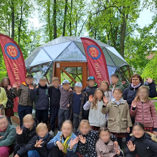 Vaikų grupė su dviem mokytojomis pozuoja prie stiklinio kupolo lauko edukacijoje. Visi stovi ar pritūpę, mojuoja į kamerą ir šypsosi. Fone – žaluma, du raudoni stendai su logotipu ir užrašais. Aplinka – pavasariškai žalias parkas.