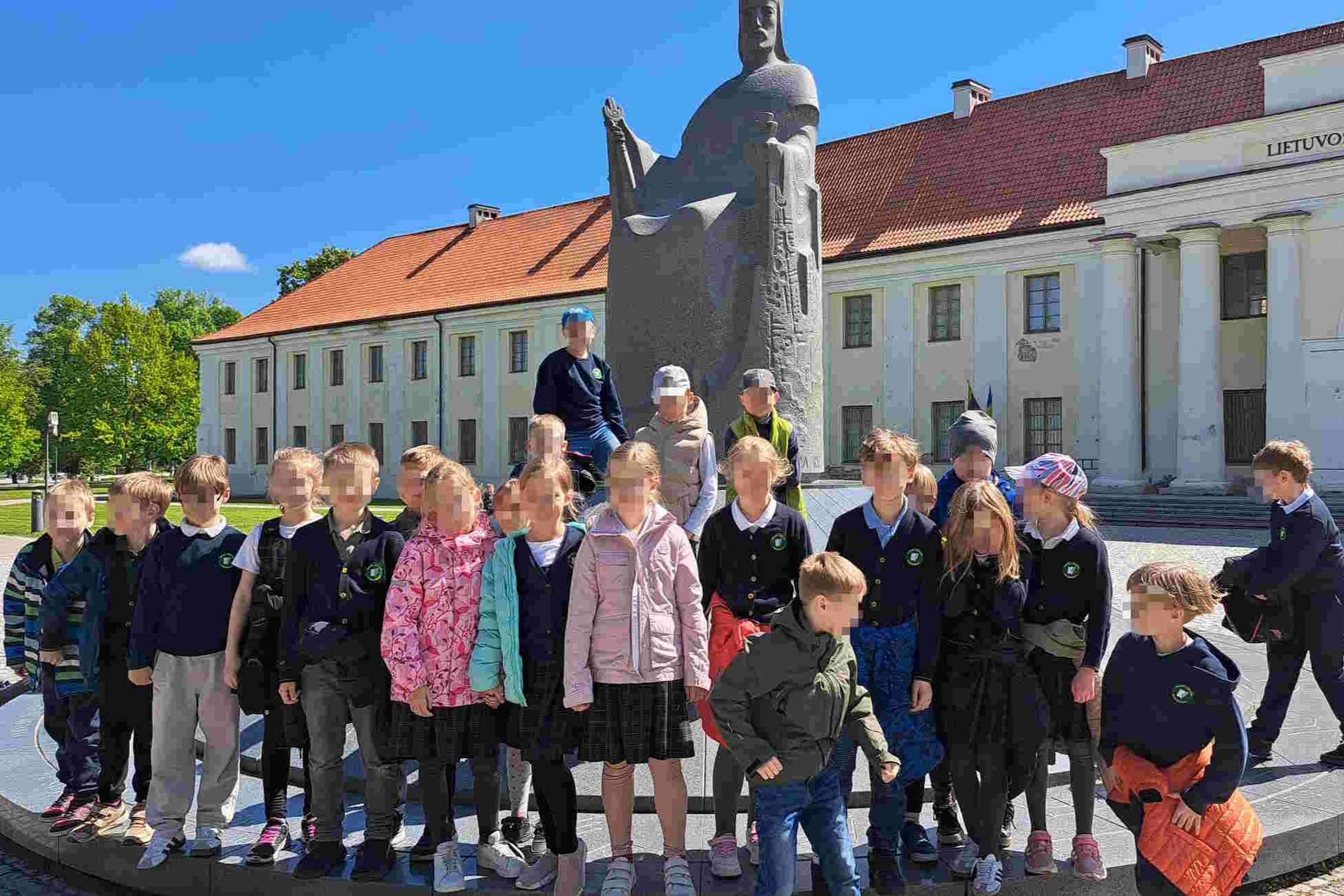 Grupė pradinių klasių mokinių pozuoja prie karaliaus Mindaugo paminklo Vilniuje, Lietuvos nacionalinio muziejaus fone. Saulėta diena, vaikai dėvi mokyklines uniformas ir spalvingus viršutinius drabužius.