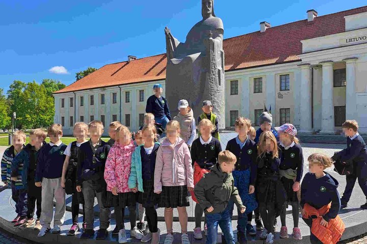 Grupė pradinių klasių mokinių pozuoja prie karaliaus Mindaugo paminklo Vilniuje, Lietuvos nacionalinio muziejaus fone. Saulėta diena, vaikai dėvi mokyklines uniformas ir spalvingus viršutinius drabužius.