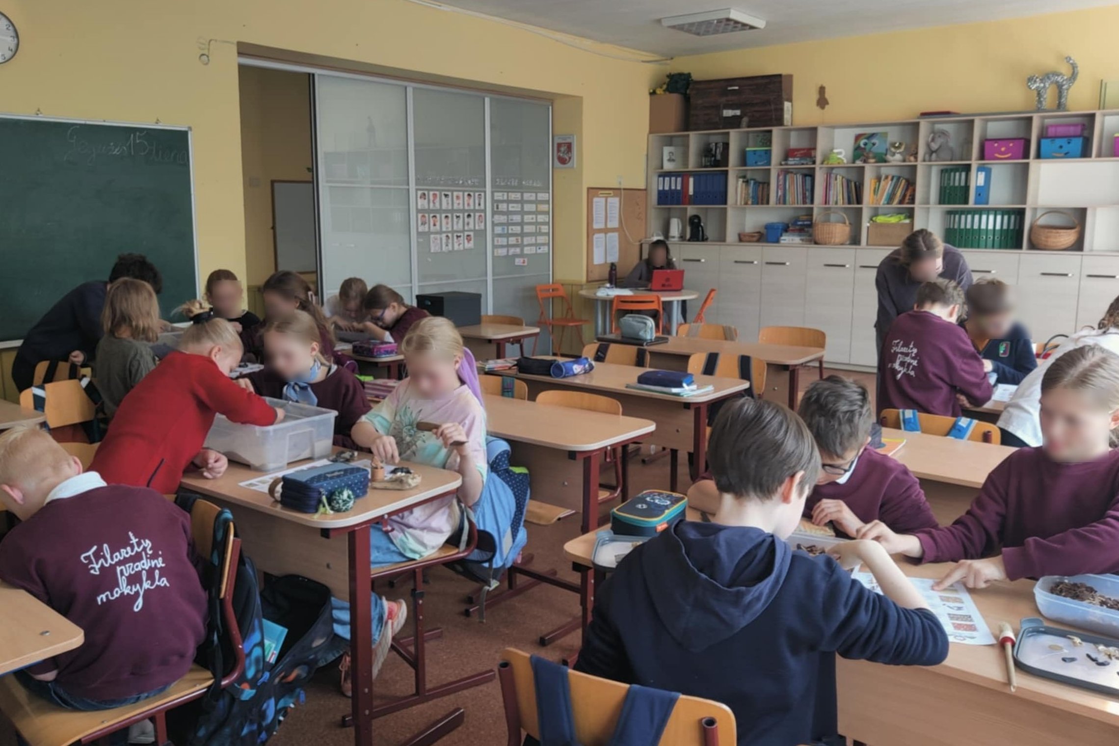 Klasės aplinkoje vaikai dirba poromis ir grupėmis prie stalų, užsiima edukacine veikla su įvairiomis medžiagomis, tai archeologinė edukacija. Ant kai kurių vaikų uniformų matyti užrašas „Filaretų pradinė mokykla“. Klasėje yra lentyna su knygomis ir priemo