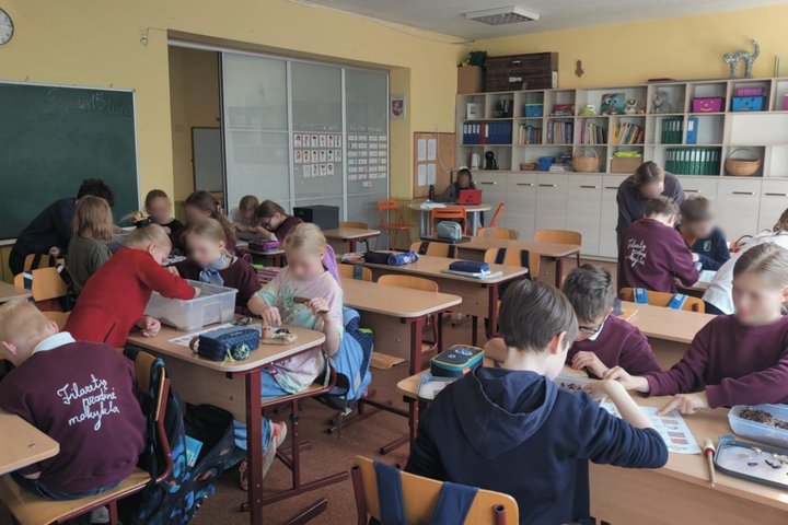 Klasės aplinkoje vaikai dirba poromis ir grupėmis prie stalų, užsiima edukacine veikla su įvairiomis medžiagomis, tai archeologinė edukacija. Ant kai kurių vaikų uniformų matyti užrašas „Filaretų pradinė mokykla“. Klasėje yra lentyna su knygomis ir priemo