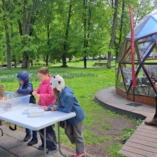 Vaikai stovi prie lauko stalo ir dalyvauja edukacinėje veikloje, stato savo piliakalnį ir pilį ant jo. Veiksmas vyksta žalioje parko aplinkoje šalia stiklinio kupolo, pažymėto raudona „Vilniaus pilys“ vėliava.