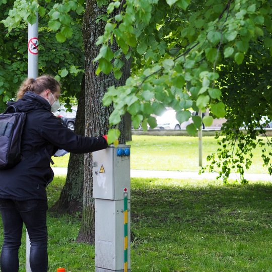 Moteris su kuprine ir apsaugine kauke atlieka veiksmą prie elektros skydinės parke. Aplink žali medžiai, žolė ir automobilių stovėjimo aikštelė fone.