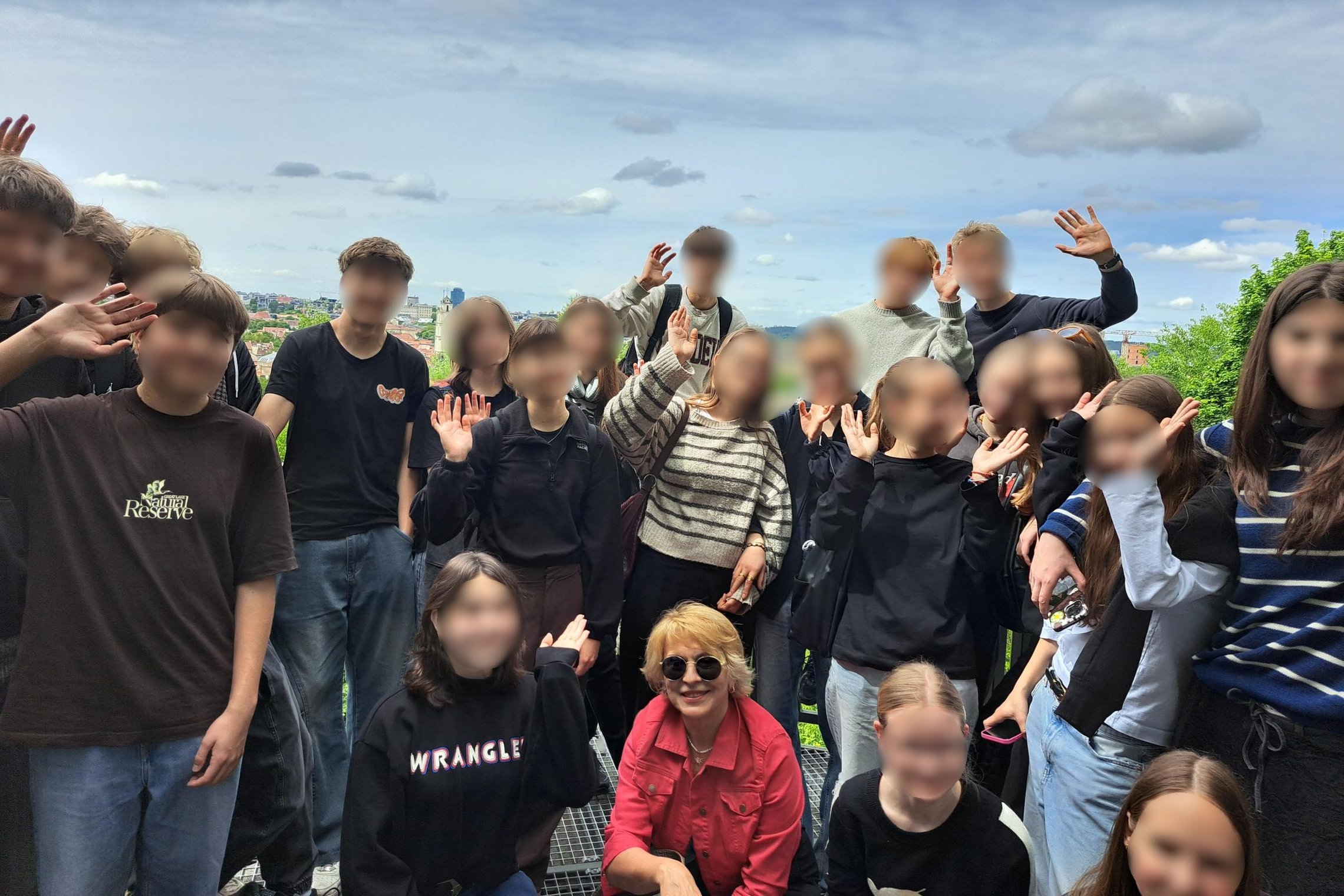 Nuotraukoje – linksma ir pozityvi jaunuolių grupė, greičiausiai moksleiviai su mokytoja, susibūrę bendram kadrui lauke. Jie šypsosi, mojuoja ir atrodo gerai nusiteikę. Priekyje, centre, matyti mokytoja su raudonu švarkeliu ir akiniais nuo saulės, o už jos