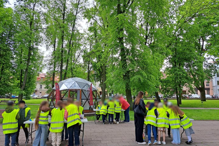 Nuotraukoje matyti grupė vaikų su ryškiaspalvėmis geltonomis liemenėmis, dalyvaujantys veiklose lauke. Jie susibūrę prie stalų, o fone stovi kupolo formos statinys – tai edukacinė erdvė, tikėtina, Vilniaus pilių kupolas. Aplink daug žalumos – vešlus parka
