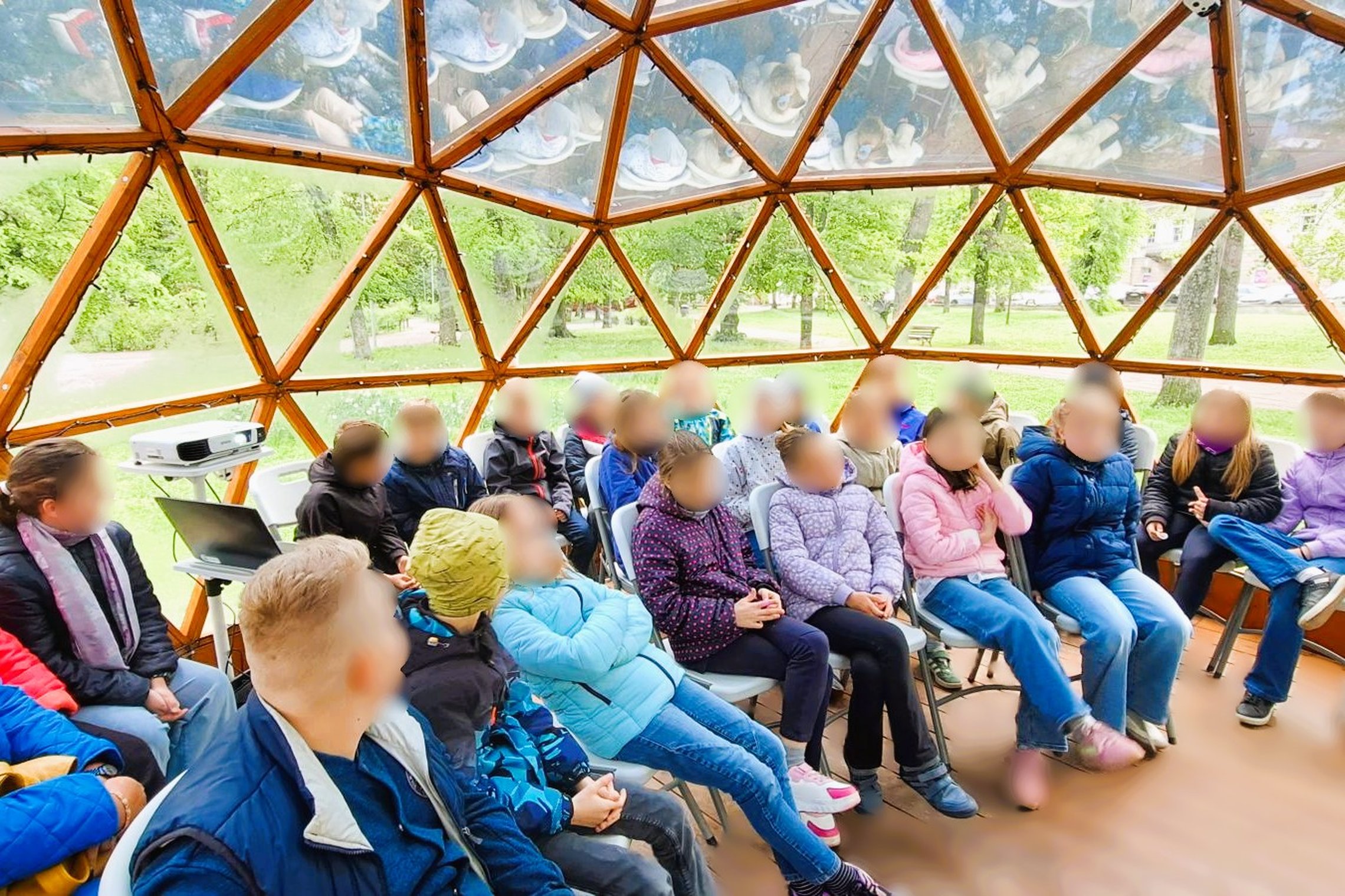Nuotraukoje – grupė vaikų, greičiausiai pradinių klasių mokiniai, susirinkę į edukacinį užsiėmimą kupolo formos pavėsinėje lauke. Jie dėvi striukes ir šiltesnius drabužius, o tai rodo, kad užsiėmimas vyksta vėsesniu metų laiku. Šalia matyti projektorius i