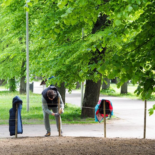 Vyras lenkiasi prie medinio kuoliuko ir tvirtina virvę pažymėtame žemės plote. Aplink žali medžiai ir pasivaikščiojimų takas parke.