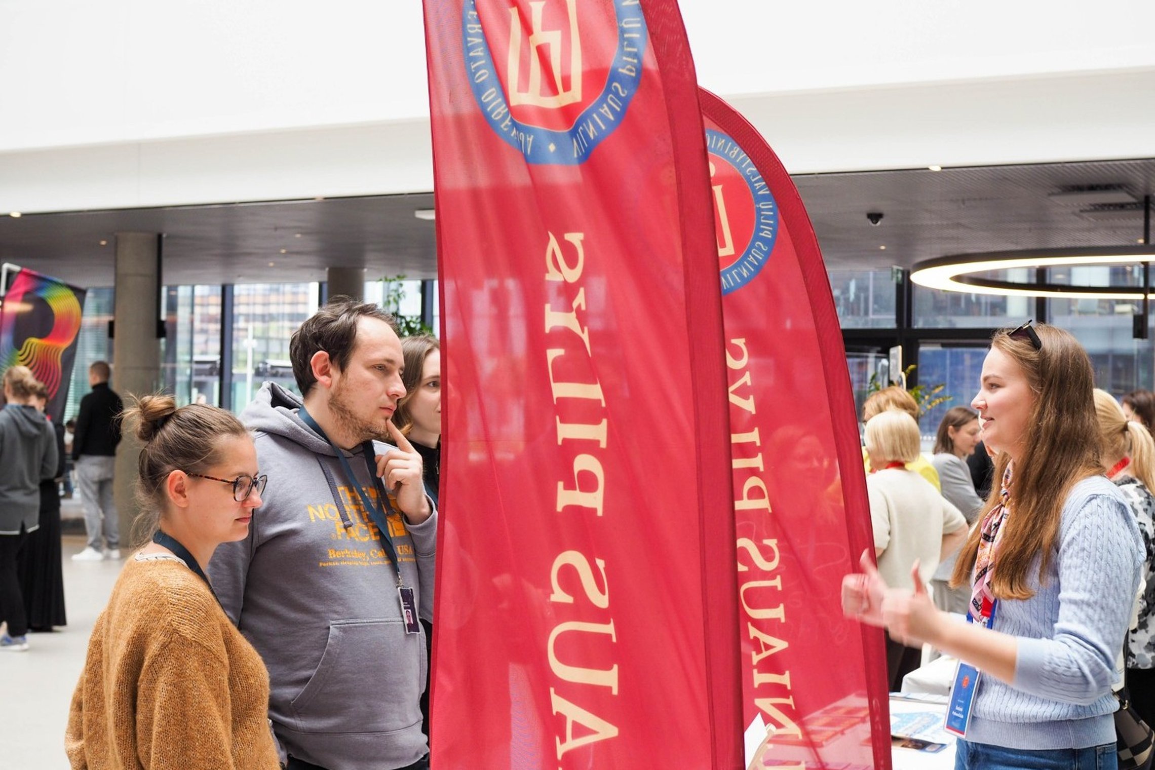 Grupė žmonių bendrauja prie informacinio stendo, pažymėto raudonomis vėliavomis su užrašu „Vilniaus pilys“. Jauna moteris dešinėje, dėvinti šviesiai mėlyną megztinį ir turinti lankytojo kortelę, paaiškina informaciją trims klausytojams. Vienas iš jų, vyra
