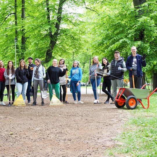 Grupė žmonių stovi parke po talkos, rankose laikydami kastuvus, grėblius ir šluotas. Visi šypsosi, fone žaliuoja medžiai ir stovi karutis.