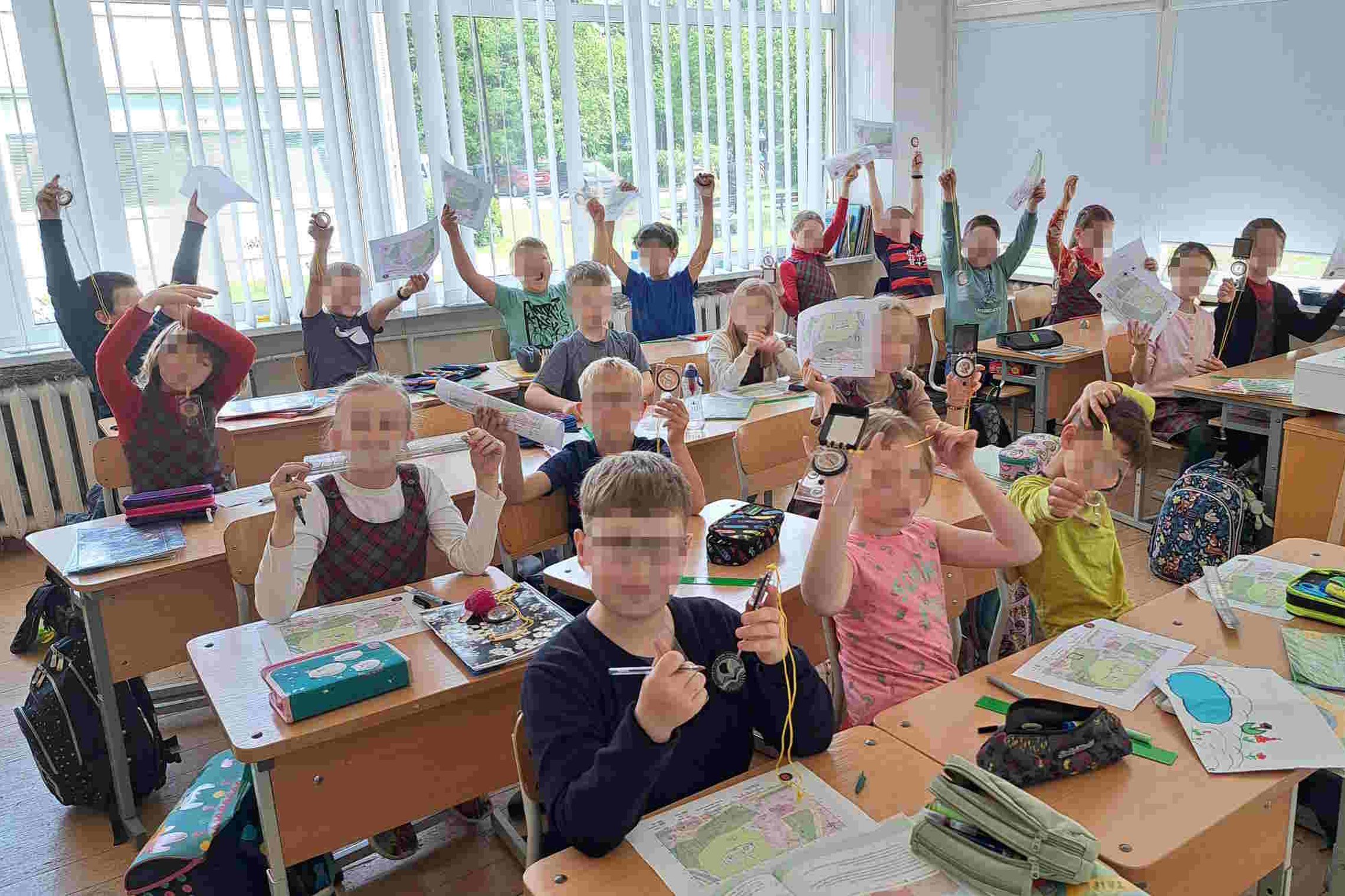 Pradinukai klasėje džiaugsmingai kelia į viršų kompasus ir žemėlapius, dalyvaudami edukacijoje. Ant stalų matyti Vilniaus pilių teritorijos planai ir mokymosi priemonės.