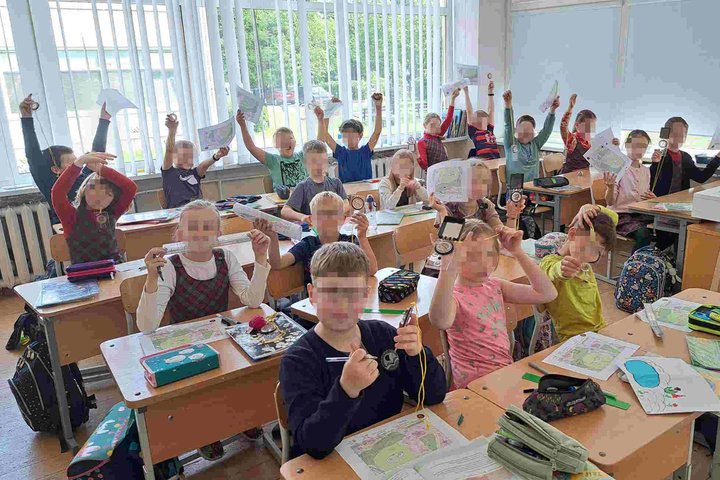 Pradinukai klasėje džiaugsmingai kelia į viršų kompasus ir žemėlapius, dalyvaudami edukacijoje. Ant stalų matyti Vilniaus pilių teritorijos planai ir mokymosi priemonės.