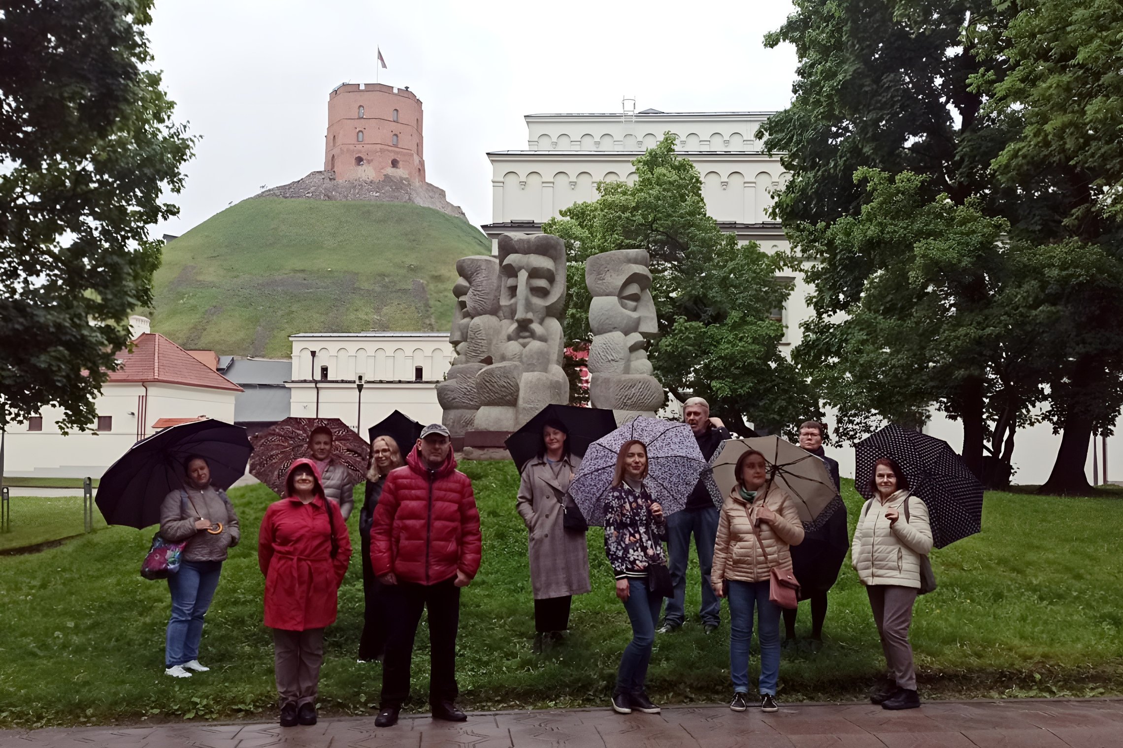Grupė žmonių stovi su skėčiais prie skulptūros „Trys Karaliai“ Vilniaus pilių teritorijoje, už jų – Gedimino pilies bokštas ant kalno. Aplink – žali medžiai ir istorinių pastatų fasadai. Nors lyja, visi šypsosi ir dalyvauja ekskursijoje.