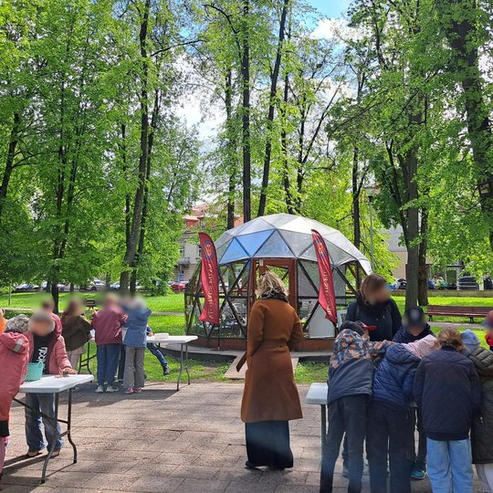 Vilniaus pilių teritorijoje, žalumos apsuptyje, vaikai dalyvauja edukacinėse veiklose prie kelių stalų lauke. Fone – stiklinis kupolas su raudonomis vėliavomis, pažymėtomis užrašu „Vilniaus pilys“.