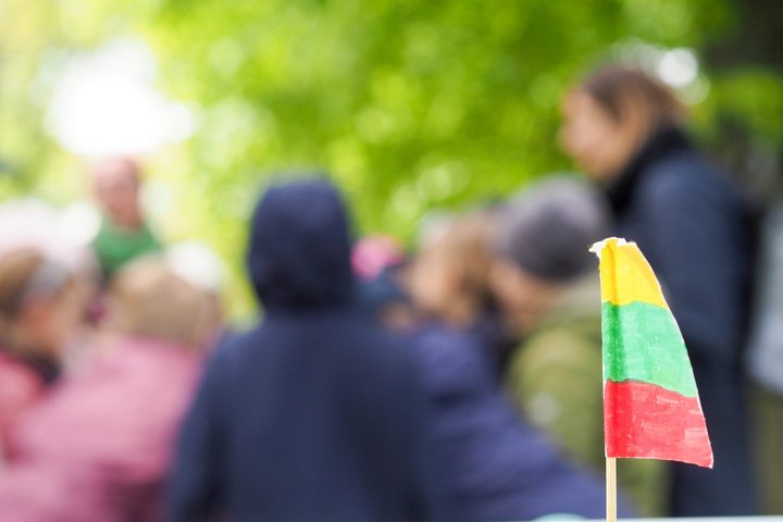 Priekyje – rankomis piešta Lietuvos vėliavėlė. Fone neryškiai matomi vaikai, pasinėrę į veiklą lauke, tikėtina – sprendžia užduotis ar ieško užuominų.