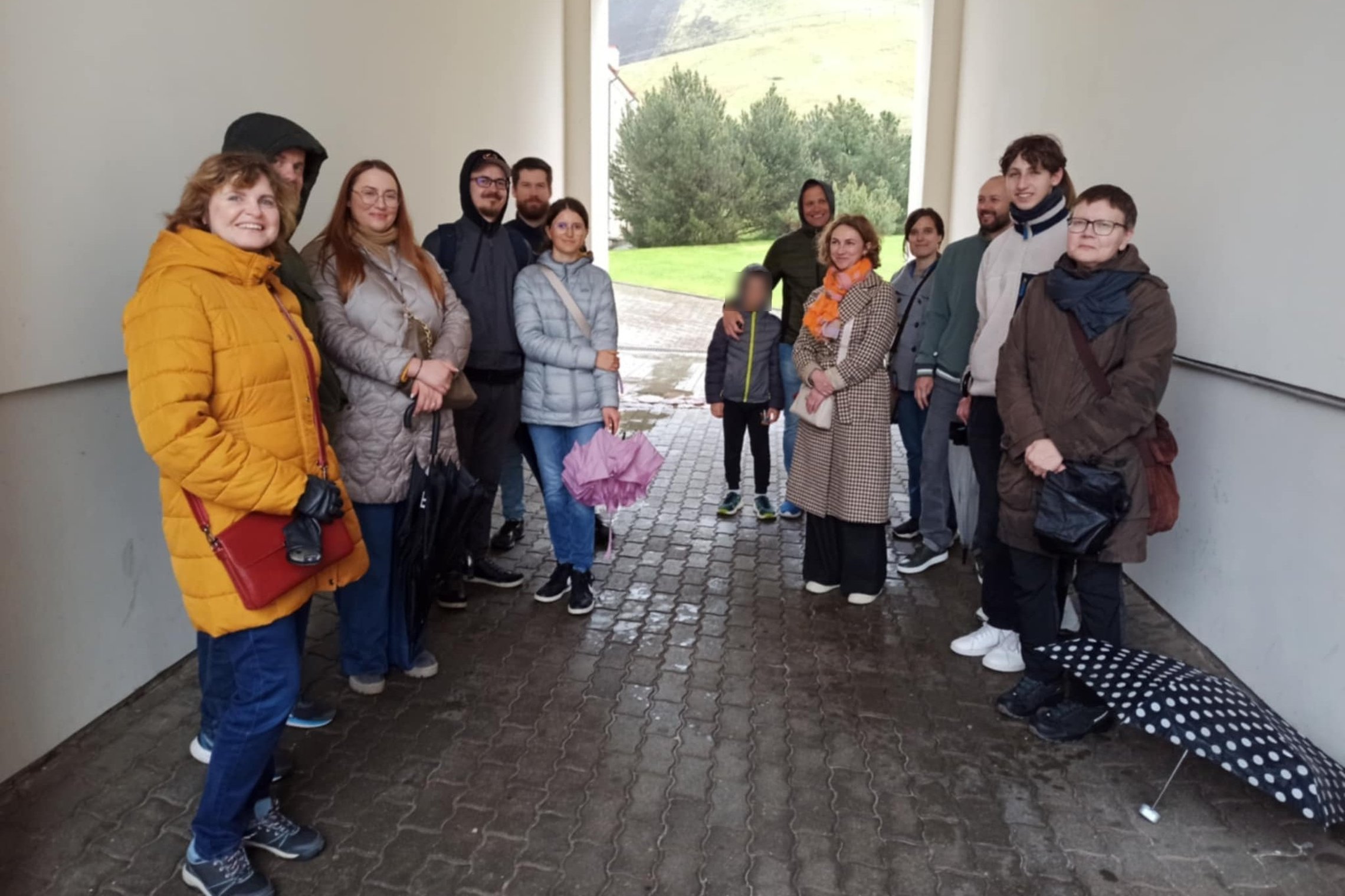 Grupė suaugusiųjų ir vaikas stovi arkadoje po stogu, apsisaugoję nuo lietaus. Dauguma vilki striukes, kai kurie rankose laiko skėčius, vienas skėtis guli ant žemės. Fone matyti žolė, medžiai ir šlaitas su Gedimino kalno dalimi. Nuotrauka daryta Vilniaus p