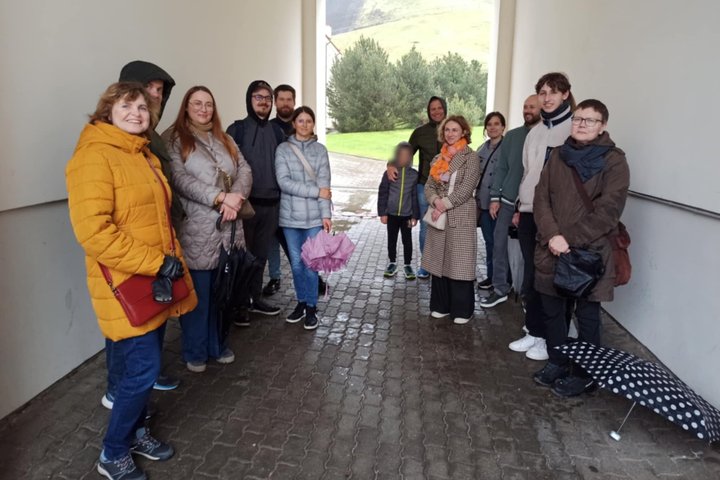 Grupė suaugusiųjų ir vaikas stovi arkadoje po stogu, apsisaugoję nuo lietaus. Dauguma vilki striukes, kai kurie rankose laiko skėčius, vienas skėtis guli ant žemės. Fone matyti žolė, medžiai ir šlaitas su Gedimino kalno dalimi. Nuotrauka daryta Vilniaus p