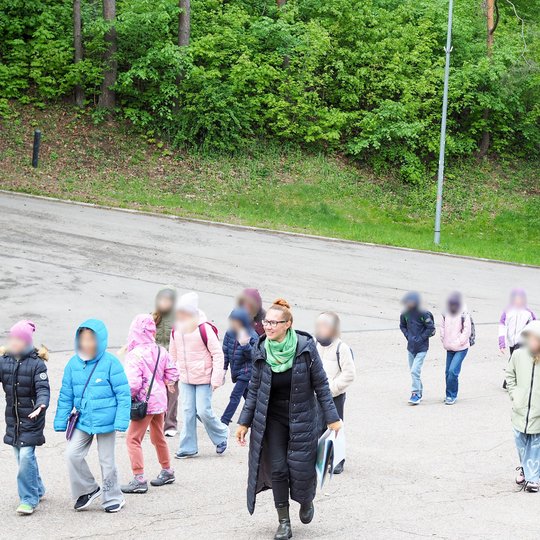 Vaikai su lydinčia edukatore eina taku šalia miškingo šlaito. Jie apsirengę šiltais drabužiais, o oras atrodo vėsus, bet šviesus.