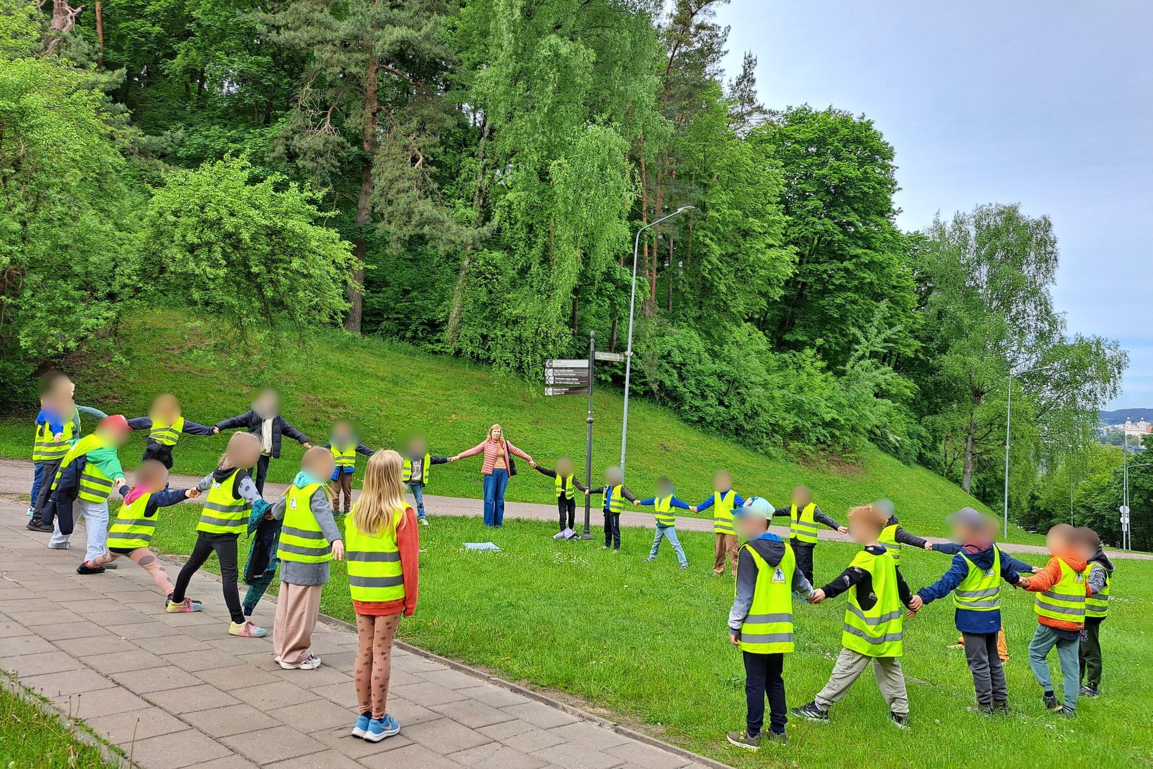 Nuotraukoje užfiksuota akimirka iš edukacinės išvykos gamtoje. Žalia, medžiais apaugusi kalva rodo, kad veiksmas vyksta gamtinėje aplinkoje, greičiausiai netoli Kreivosios pilies vietos Vilniuje. Pirmame plane – didelė grupė vaikų, vilkinčių ryškias gelt