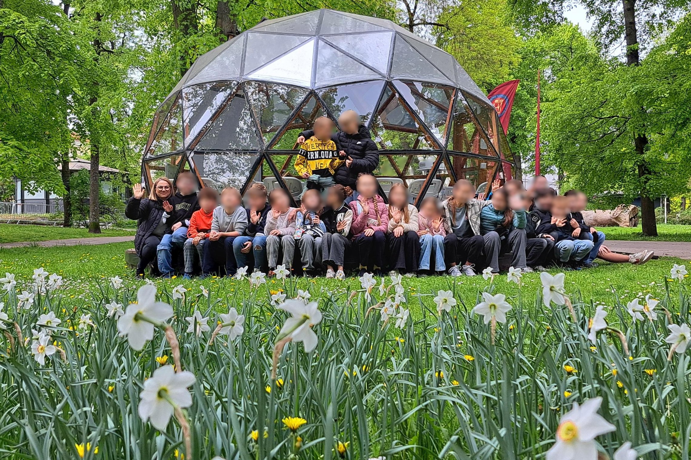 Grupė vaikų su mokytojais pozuoja nuotraukai žaliuojančiame parke prie stiklinio kupolo formos statinio. Priekyje žydi balti narcizai, o fone matyti medžiai ir pavasariškai sodri gamta. Visi sėdi ar stovi šypsodamiesi, kai kurie mojuoja į kamerą.