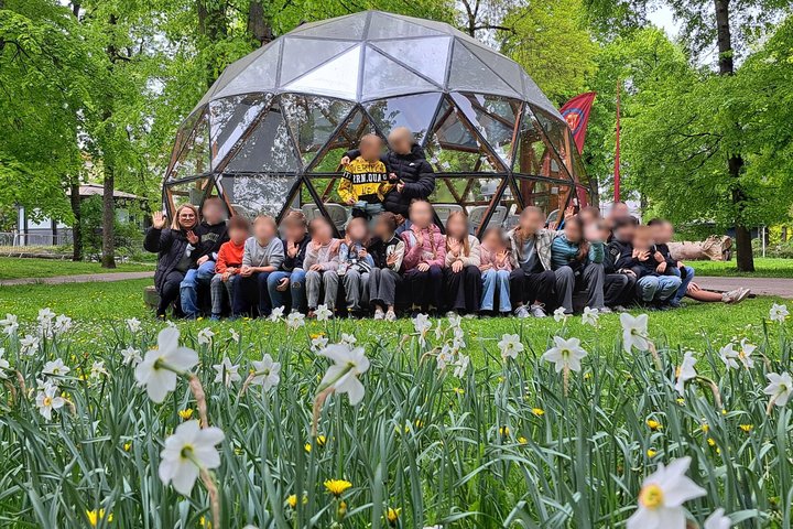 Grupė vaikų su mokytojais pozuoja nuotraukai žaliuojančiame parke prie stiklinio kupolo formos statinio. Priekyje žydi balti narcizai, o fone matyti medžiai ir pavasariškai sodri gamta. Visi sėdi ar stovi šypsodamiesi, kai kurie mojuoja į kamerą.