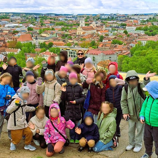 Vaikai ir mokytoja pozuoja Trijų Kryžių kalno apžvalgos aikštelėje, už jų – platus Vilniaus senamiesčio panoraminis vaizdas su raudonais stogais ir bažnyčių bokštais.