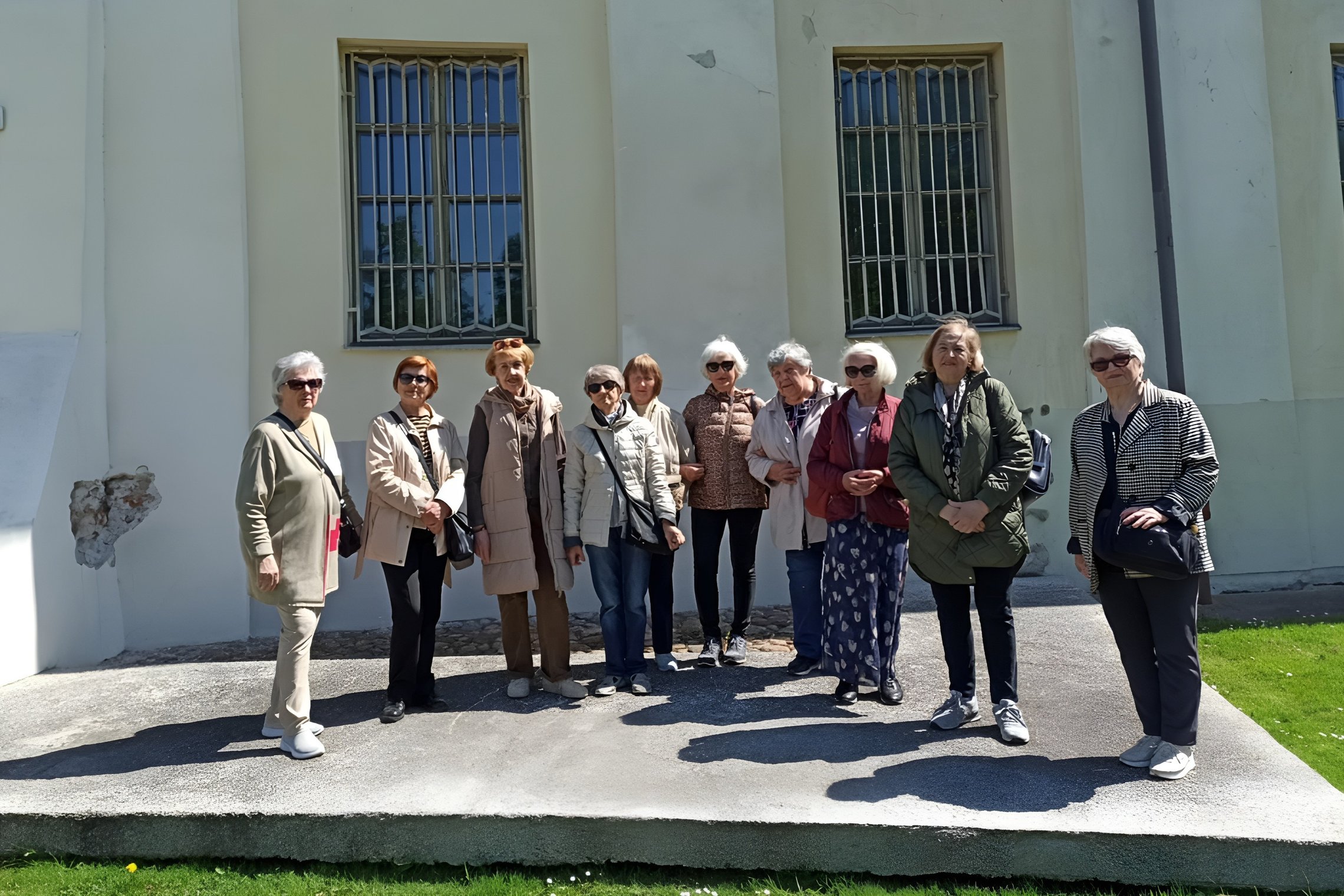 Grupė senjorių stovi lauke prie šviesios sienos su dviem langais, uždengtais grotomis. Moterys vilki švarkus ir striukes, pozuoja saulėtą dieną, žolė pirmame plane, šešėliai krenta ant grindinio. Tai Trečiojo amžiaus universiteto dalyvės ekskursijos metu.