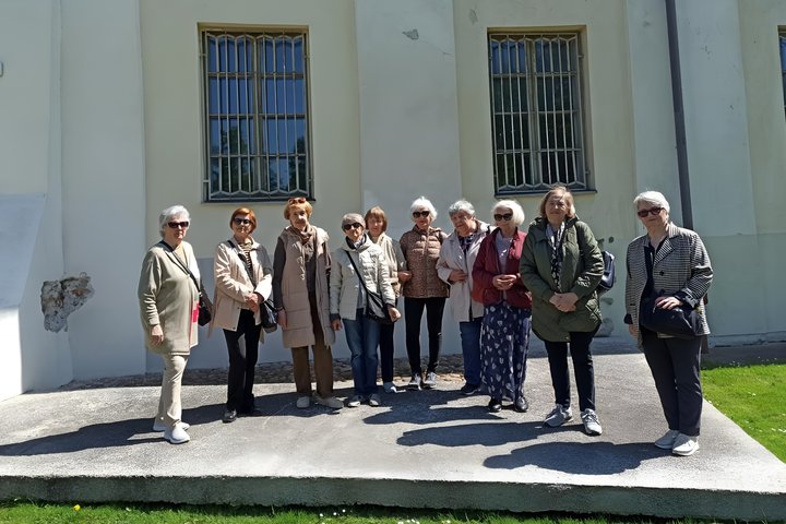 Grupė senjorių stovi lauke prie šviesios sienos su dviem langais, uždengtais grotomis. Moterys vilki švarkus ir striukes, pozuoja saulėtą dieną, žolė pirmame plane, šešėliai krenta ant grindinio. Tai Trečiojo amžiaus universiteto dalyvės ekskursijos metu.