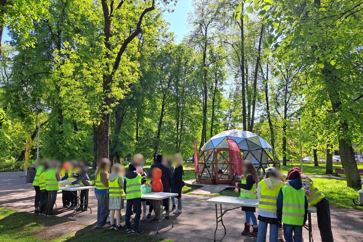 Vaizdas užfiksuoja vaikų grupę, dalyvaujančią edukaciniame užsiėmime lauke, saulėtą dieną žaliuojančiame parke. Mokiniai, vilkintys ryškias geltonas liemenes, susibūrę prie kelių stalų, kur, panašu, vyksta kūrybinė veikla. Fone matyti modernus kupolo form