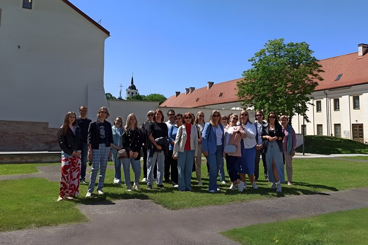 Grupė žmonių stovi žalioje vejoje prie istorinio pastato su baltomis sienomis ir raudonu čerpiniu stogu, saulėtą dieną. Fone matyti Vilniaus katedros varpinė. Tai Vilniaus pilių teritorija. Visi pozuoja bendroje nuotraukoje, apsirengę šiuolaikiškai. – kai