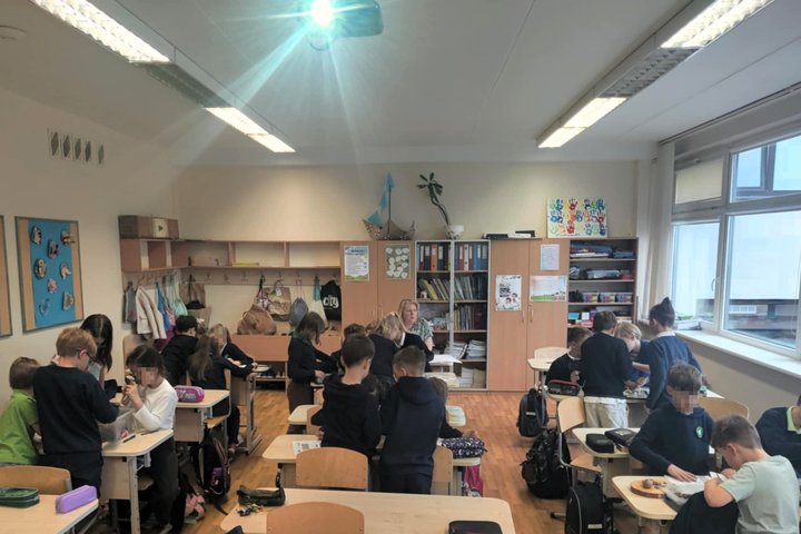 Klasėje dirba būreliai vaikų – visi susitelkę ties įvairiomis veiklomis. Vaikai stovi ar sėdi prie stalų, tiria daiktus, diskutuoja tarpusavyje. Tai pradinių klasių mokiniai, tapę archeologais per edukaciją. Fone matyti mokytoja, lentynos su knygomis ir s