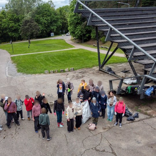 Vaikai su mokytojais stovi ratu prie lauko scenos Kalnų parke. Aplink matyti kuprinės, šiukšlių maišai ir žalios erdvės takeliai.
