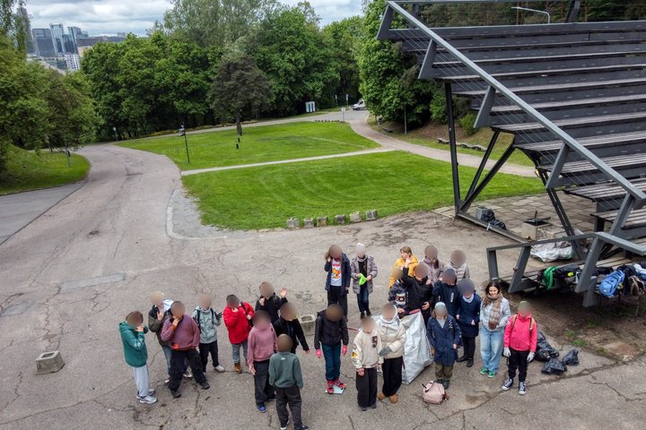 Vaikai su mokytojais stovi ratu prie lauko scenos Kalnų parke. Aplink matyti kuprinės, šiukšlių maišai ir žalios erdvės takeliai.