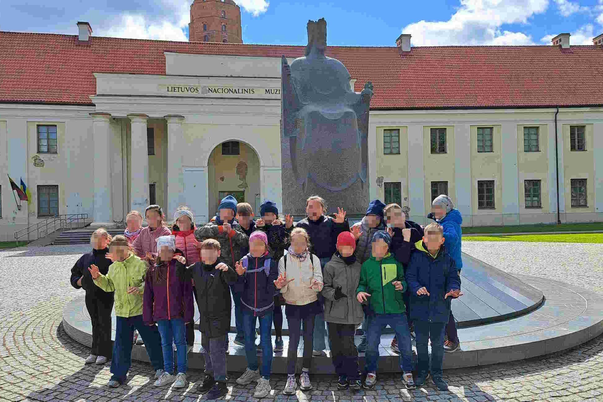 Grupė vaikų pozuoja prie Mindaugo sosto paminklo aikštėje priešais Lietuvos nacionalinį muziejų Vilniuje. Už jų matyti muziejaus pastatas su Gedimino pilies bokštu fone.