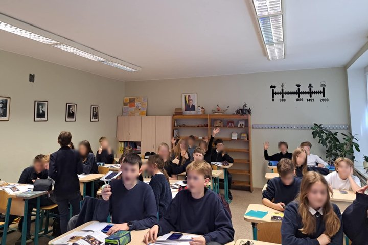 Klasėje sėdi keliolika mokinių, kai kurie pakėlę rankas ar žiūri į mokytoją. Ant stalų – vadovėliai, sąsiuviniai, kai kurie laiko telefonus. Fone matyti lentyna, spintos, ant sienos pakabinti istoriniai portretai ir laiko juosta su reikšmingomis datomis. 