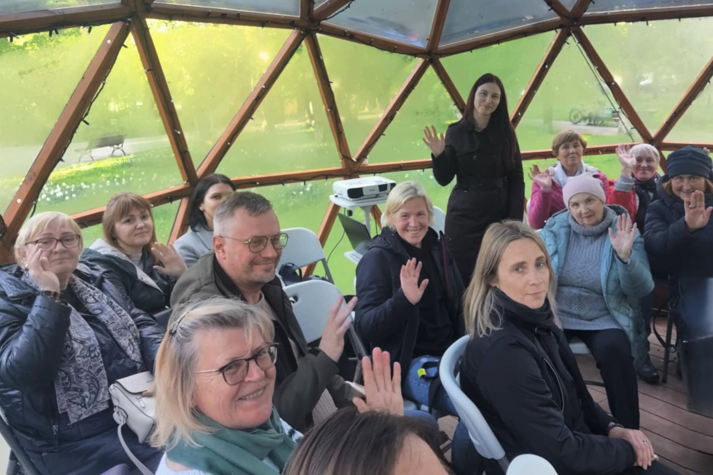 Grupė suaugusių žmonių sėdi puslankiu Vilniaus pilių kupolo viduje, medinėje struktūroje su permatomomis sienomis. Dauguma dalyvių šypsosi ir mojuoja į kamerą. Fone matomas projektorius ant trikojės lentynos, o pro kupolo langus matyti žalias parkas ir su