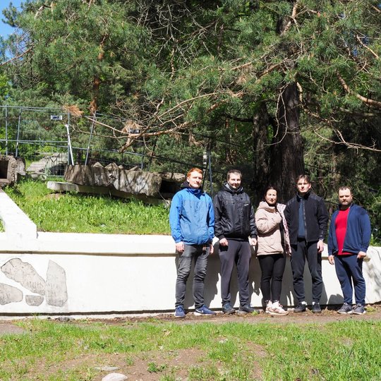 Šeši žmonės po talkos pozuoja prie balto pamato, už kurio stovi medžiai ir tvoromis aptvertos istorinės liekanos. Kairėje fone matyti Trys kryžiai – vienas iš Vilniaus simbolių.