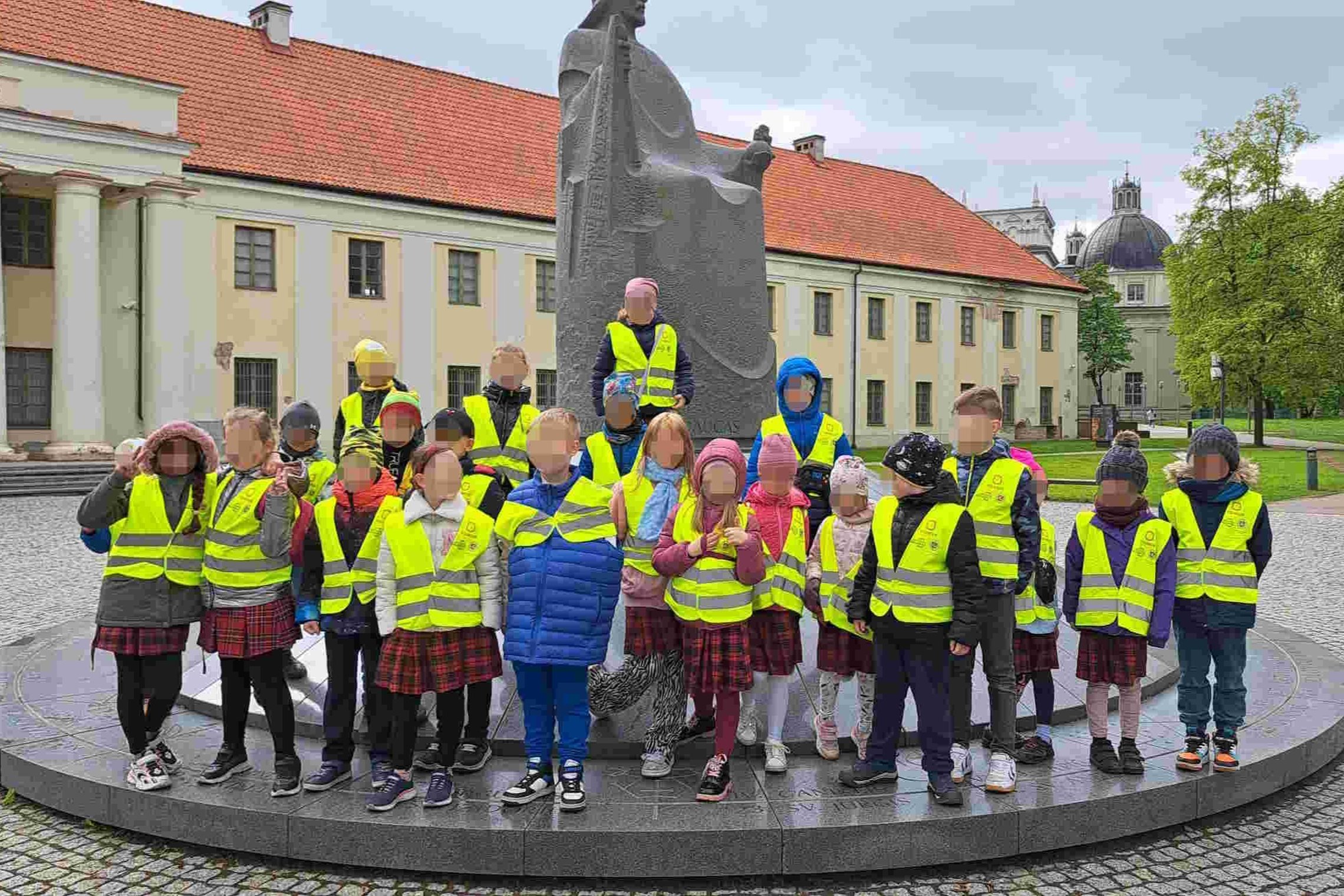 Grupė pradinių klasių mokinių su ryškiais geltonais atšvaitiniais liemenėmis pozuoja nuotraukai prie Mindaugo paminklo Vilniuje. Vaikai stovi ratu aplink granito paminklą, vaizduojantį sėdintį karalių, o fone matyti pastatas su raudonu stogu ir Vilniaus k