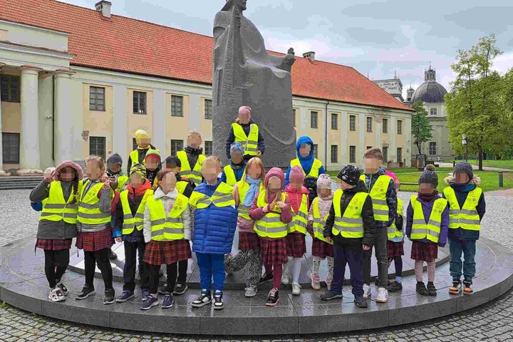 Grupė pradinių klasių mokinių su ryškiais geltonais atšvaitiniais liemenėmis pozuoja nuotraukai prie Mindaugo paminklo Vilniuje. Vaikai stovi ratu aplink granito paminklą, vaizduojantį sėdintį karalių, o fone matyti pastatas su raudonu stogu ir Vilniaus k