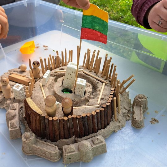 Nuotraukoje akcentuojamos vaikų sukurtos piliakalnių miniatiūros – su gynybinėmis sienomis, vartais, pastatais ir Lietuvos trispalve, iškeltą centrinėje konstrukcijos dalyje. Šie darbeliai liudija ne tik apie kūrybiškumą, bet ir apie pagarbą Lietuvos isto