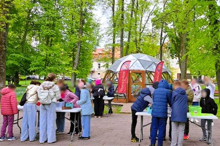Grupė vaikų dalyvauja edukacinėje veikloje lauke – jie dirba prie stalų žalioje parko aplinkoje. Fone matyti kupolo formos medinis pastatas ir raudonos vėliavos. Aplink žaliuoja medžiai, vaikams smalsiai įsitraukus į užduotis.