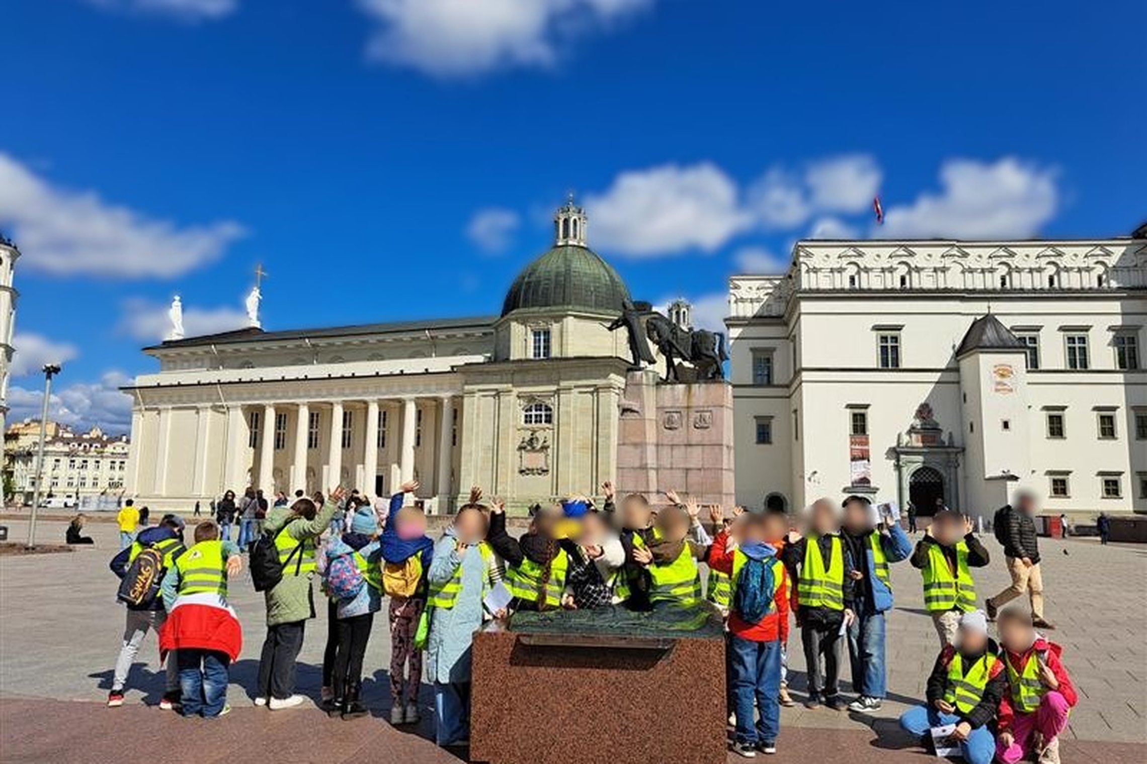 Vaikų grupė su šviesą atspindinčiomis liemenėmis stovi Katedros aikštėje Vilniuje, prie Gedimino paminklo; fone matyti Vilniaus katedra ir Valdovų rūmai, virš jų – mėlynas dangus su baltais debesimis.
